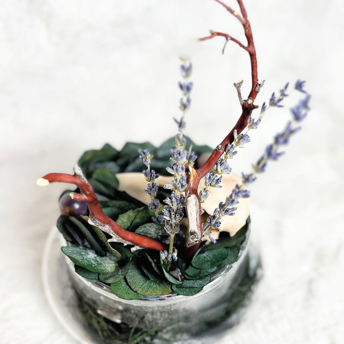A decorative dried floral arrangement in a small clear glass container, featuring green leaves, purple lavender-like sprigs, and reddish-brown branches, set against a soft white textured background.