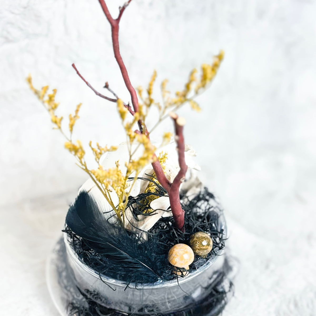 A decorative preserved egg in a clear glass container with black moss, yellow dried flowers, a reddish branch, a black feather, and two small gold-toned eggs, set against a soft white textured background.