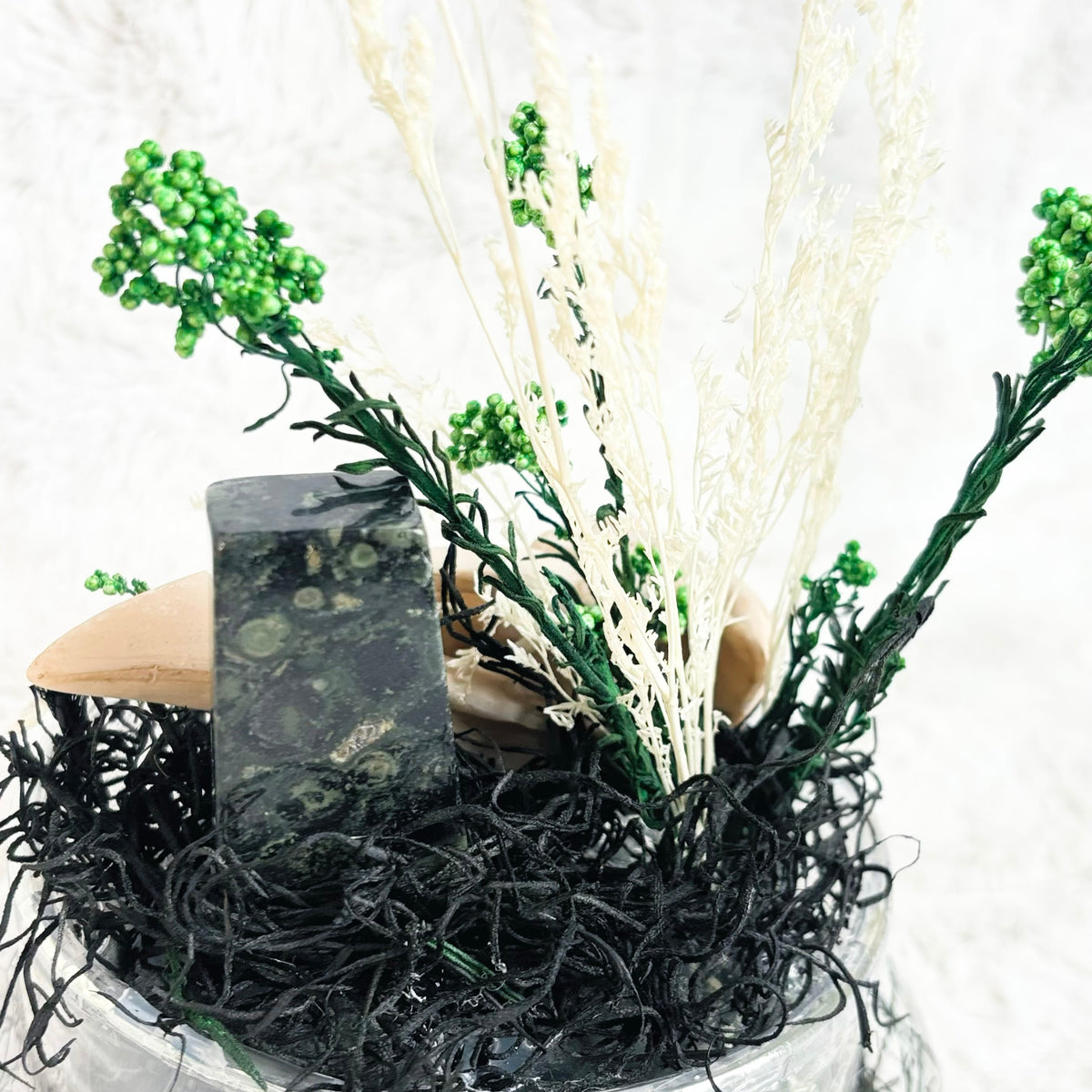 A decorative arrangement in a clear glass bowl featuring black seaweed, dried white grasses, green berry sprigs, and natural stones on a white textured background.