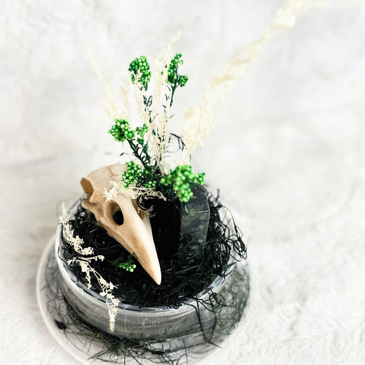 A decorative gothic-style centerpiece featuring a beige bird skull resting on a black base filled with dark shredded material, accented by small green beaded sprigs and dried white grasses, set against a soft white textured background.