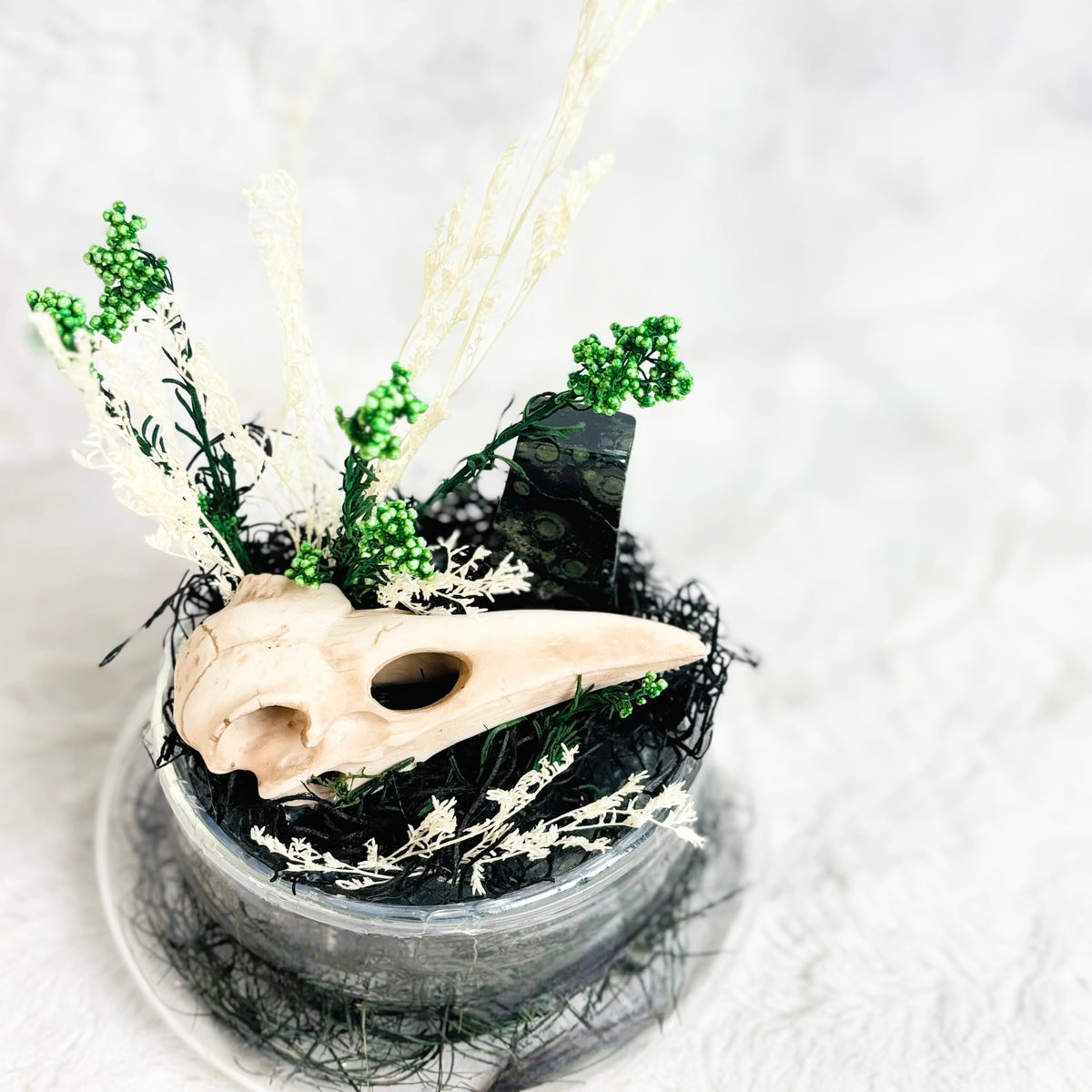A decorative Halloween centerpiece featuring a bird skull, black lace, green glittery foliage, and dried white grasses arranged in a small clear glass bowl on a white textured background.