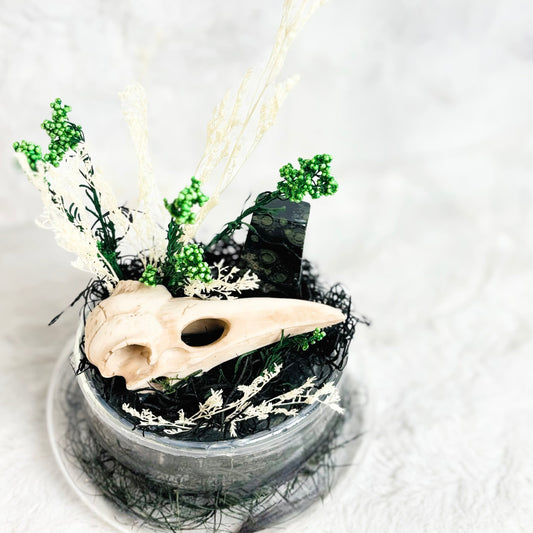 A decorative Halloween centerpiece featuring a bird skull, black lace, green glittery foliage, and dried white grasses arranged in a small clear glass bowl on a white textured background.