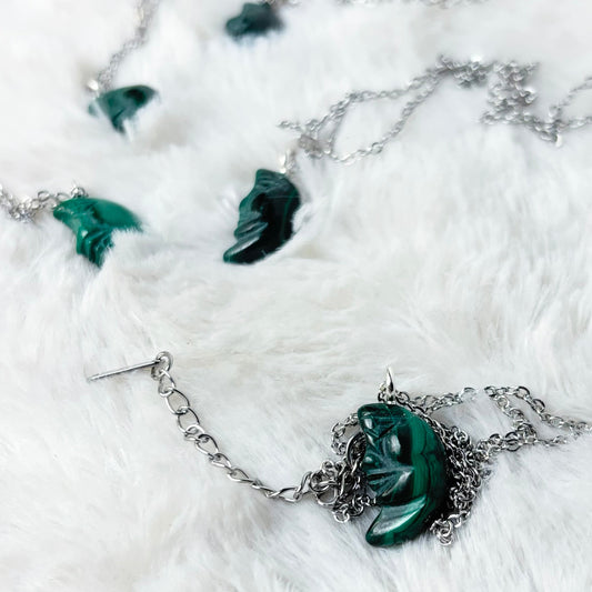 A silver-toned necklace with a carved green malachite crescent moon pendant, displayed on a white faux fur background.