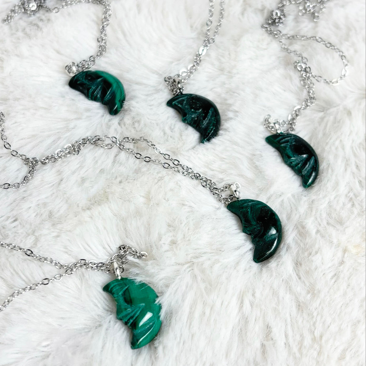 Four silver chain necklaces with polished green malachite crescent moon pendants, arranged on a white faux fur background.