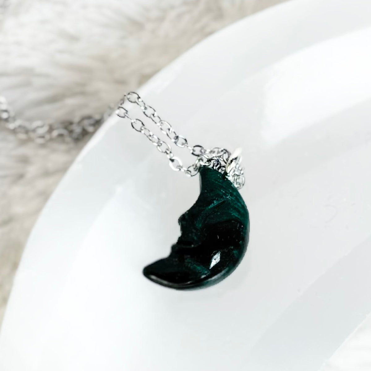 A silver-toned necklace with a crescent moon pendant containing dark green inclusions, displayed on a curved white stone holder over a white faux fur background.