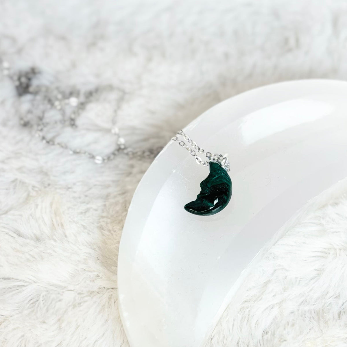 A silver necklace with a crescent moon pendant featuring a dark green stone, displayed on a white curved display stand over a white faux fur surface.