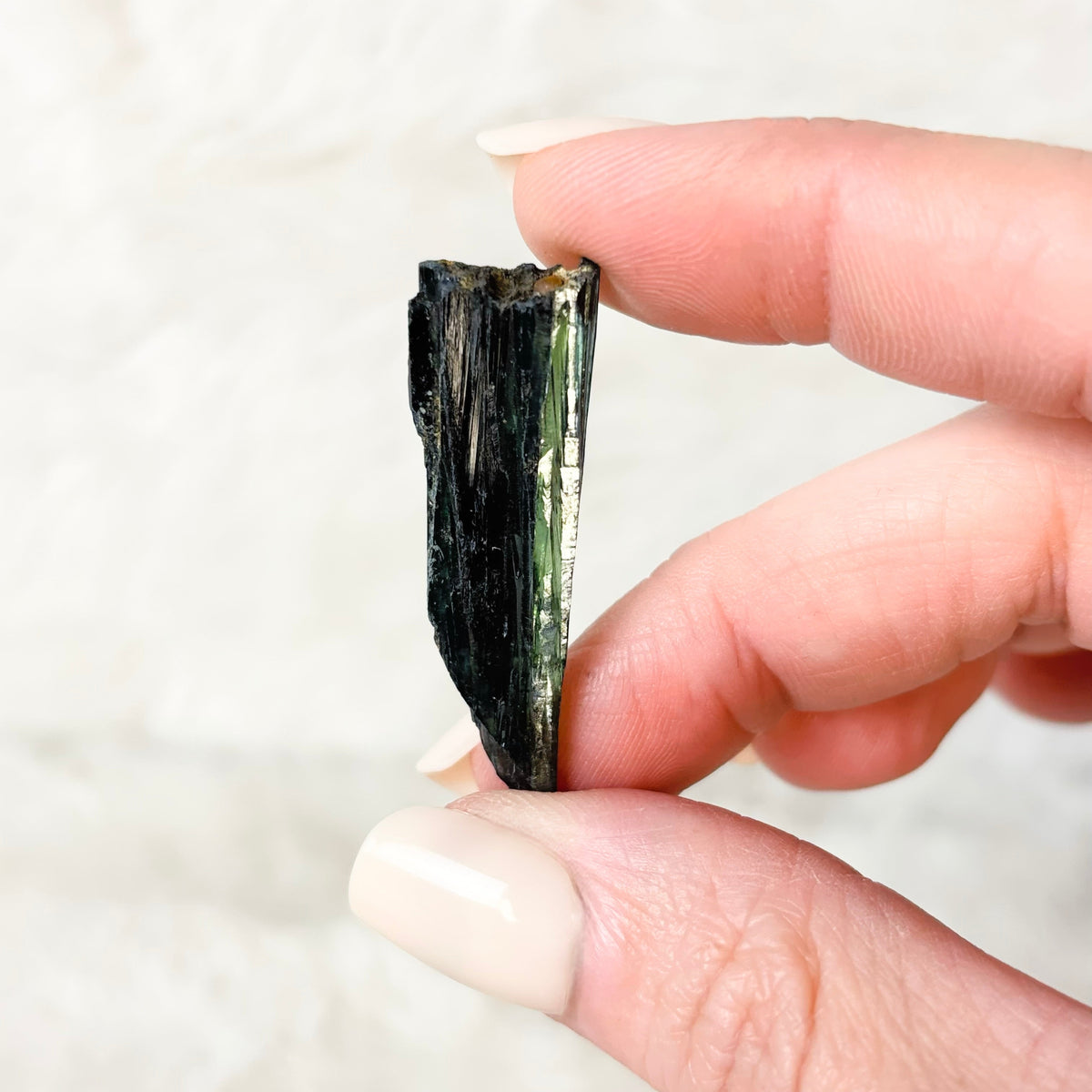 A hand with light pink manicured nails holds a small, rough, dark green to black mineral or crystal specimen against a soft, light beige textured background.