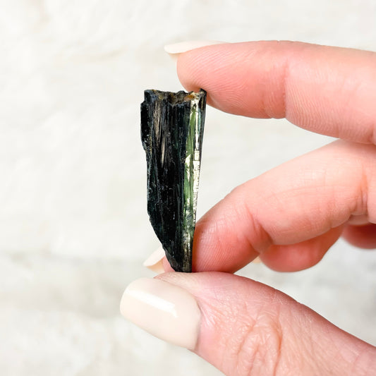 A hand with light pink manicured nails holds a small, rough, dark green to black mineral or crystal specimen against a soft, light beige textured background.