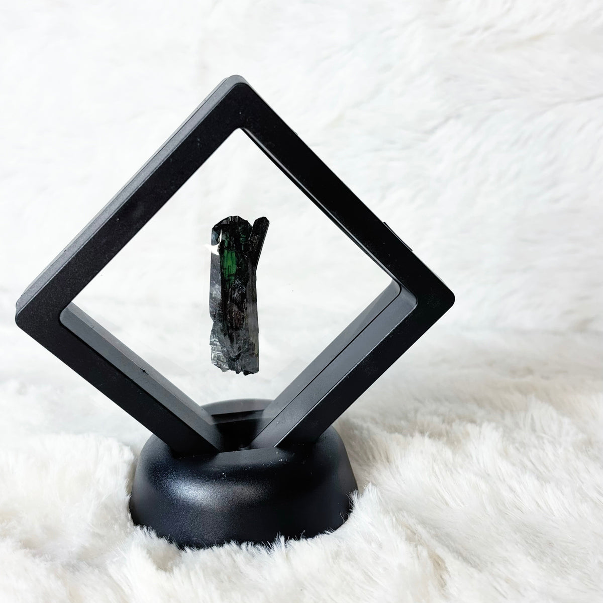 A black diamond-shaped display frame mounted on a round black base, holding a small dark mineral specimen with a green inlay, set against a white faux fur background.