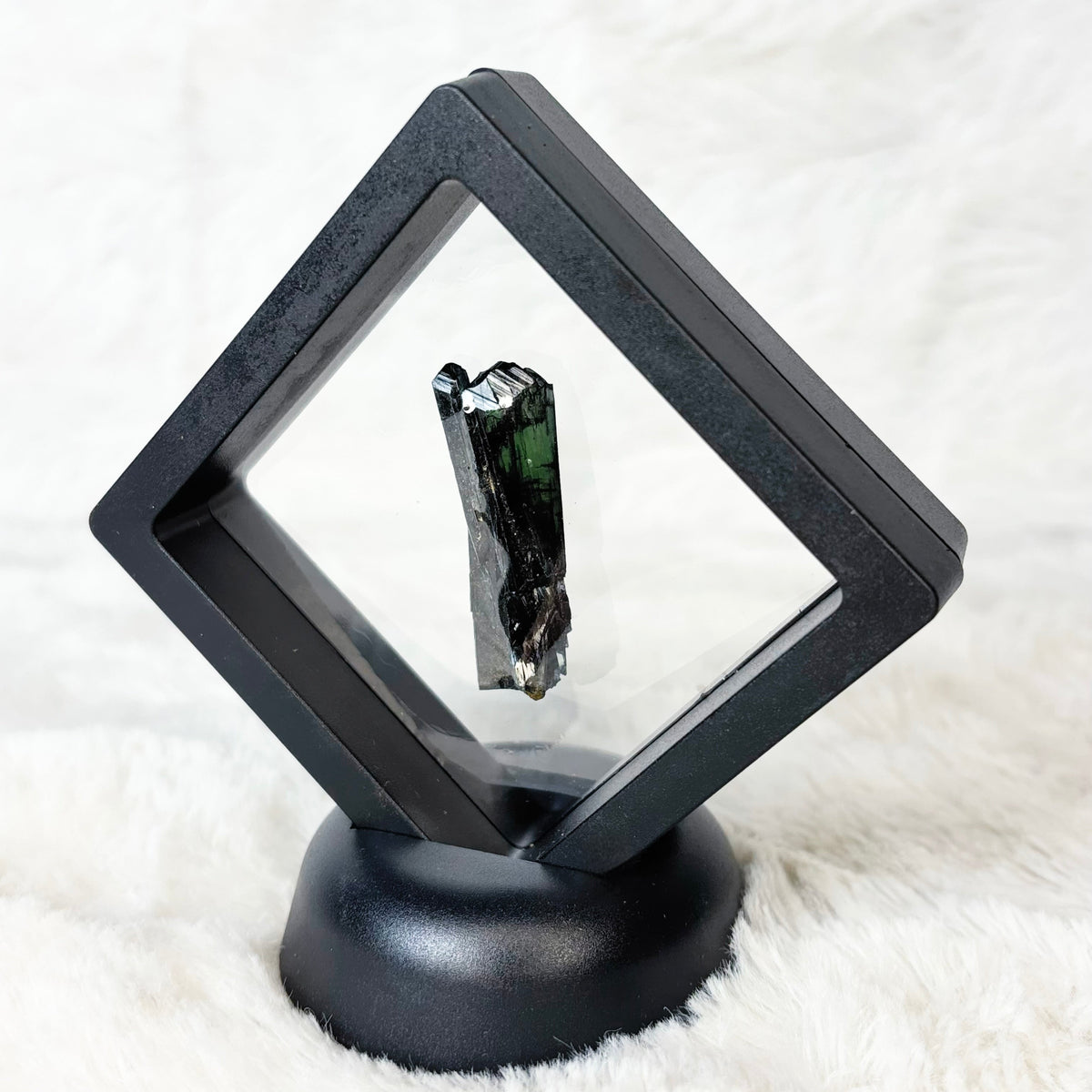 A black diamond-shaped shadow box display stand holding a small dark crystal specimen, set on a round black base against a white faux fur background.