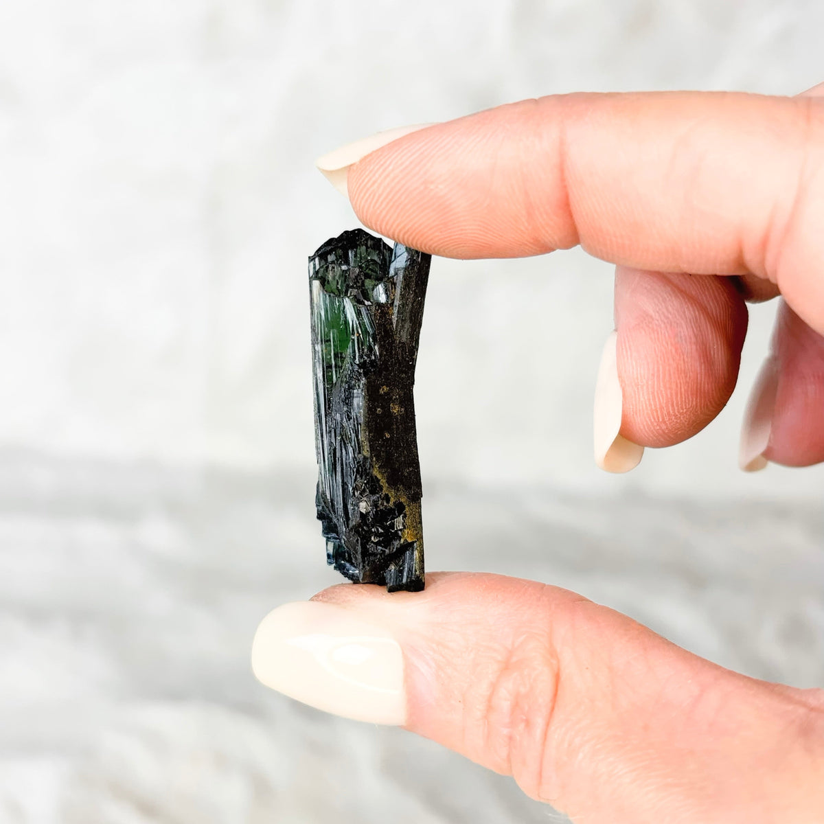 A hand with light pink manicured nails holds a small, dark green and black mineral crystal against a soft, light gray textured background.