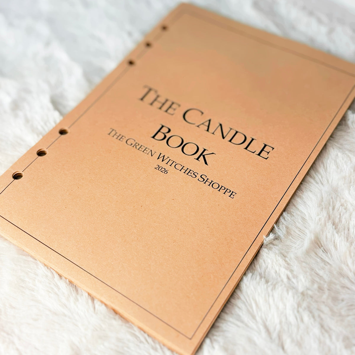 A tan leather-bound book titled 'THE CANDLE BOOK' with the subtitle 'THE GREEN WITCHES SHOPPE' and '2026' printed on the cover, resting on a white faux fur surface.