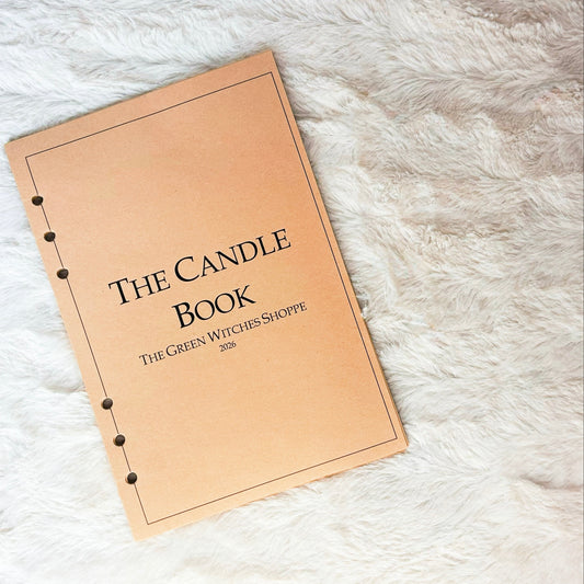 A tan spiral-bound notebook titled 'THE CANDLE BOOK' with the subtitle 'THE GREEN WITCHS SHOPPE 2026' printed on the cover, placed on a white faux fur surface.