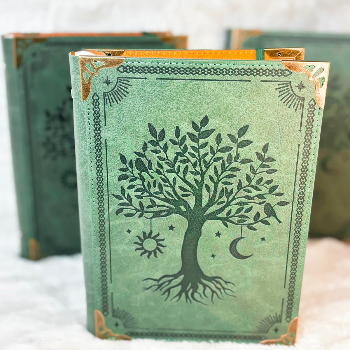 A decorative hardcover journal with a sage green cover featuring a black tree of life design and gold corner accents, standing upright with other matching journals blurred in the background.