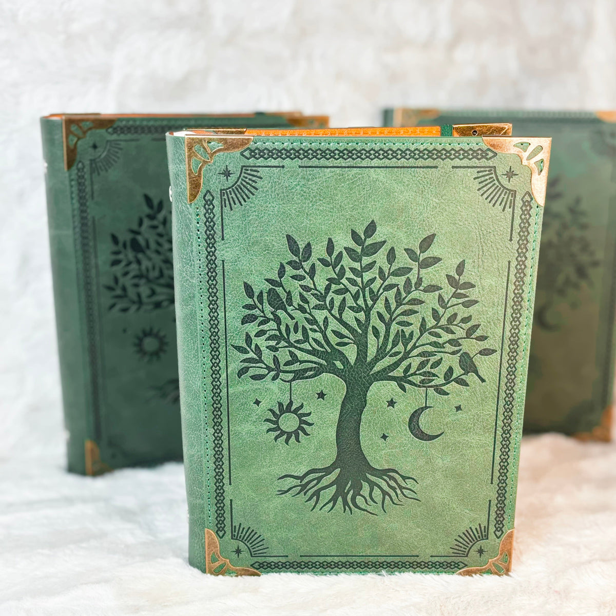 A green hardcover journal with a black tree of life design and gold corner accents, standing upright with two matching journals blurred in the background on a white textured surface.