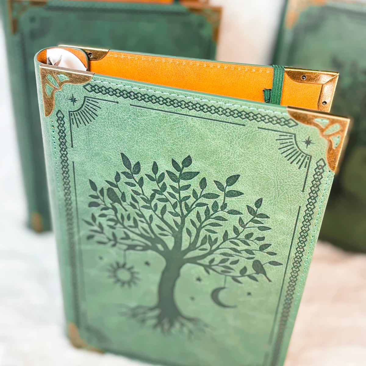 A decorative journal with a textured sage green cover featuring a black tree of life design, gold-toned corner protectors, and an orange inner page visible at the top, shown on a soft white surface.