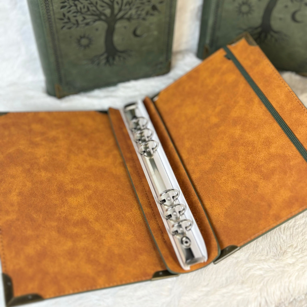 A rustic leather ring binder journal shown open, featuring a silver flute nestled inside the rings, with two closed journals displaying a tree-of-life design in the background.