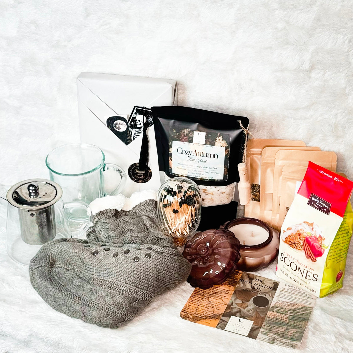 A cozy holiday gift set arranged on a white faux fur background, featuring gray cable-knit slipper socks, a glass mug and teapot with metal infuser, a bag of Cozy Autumn Bath Soak, a box of gourmet scone mix, a candle, and other warm seasonal accessories.