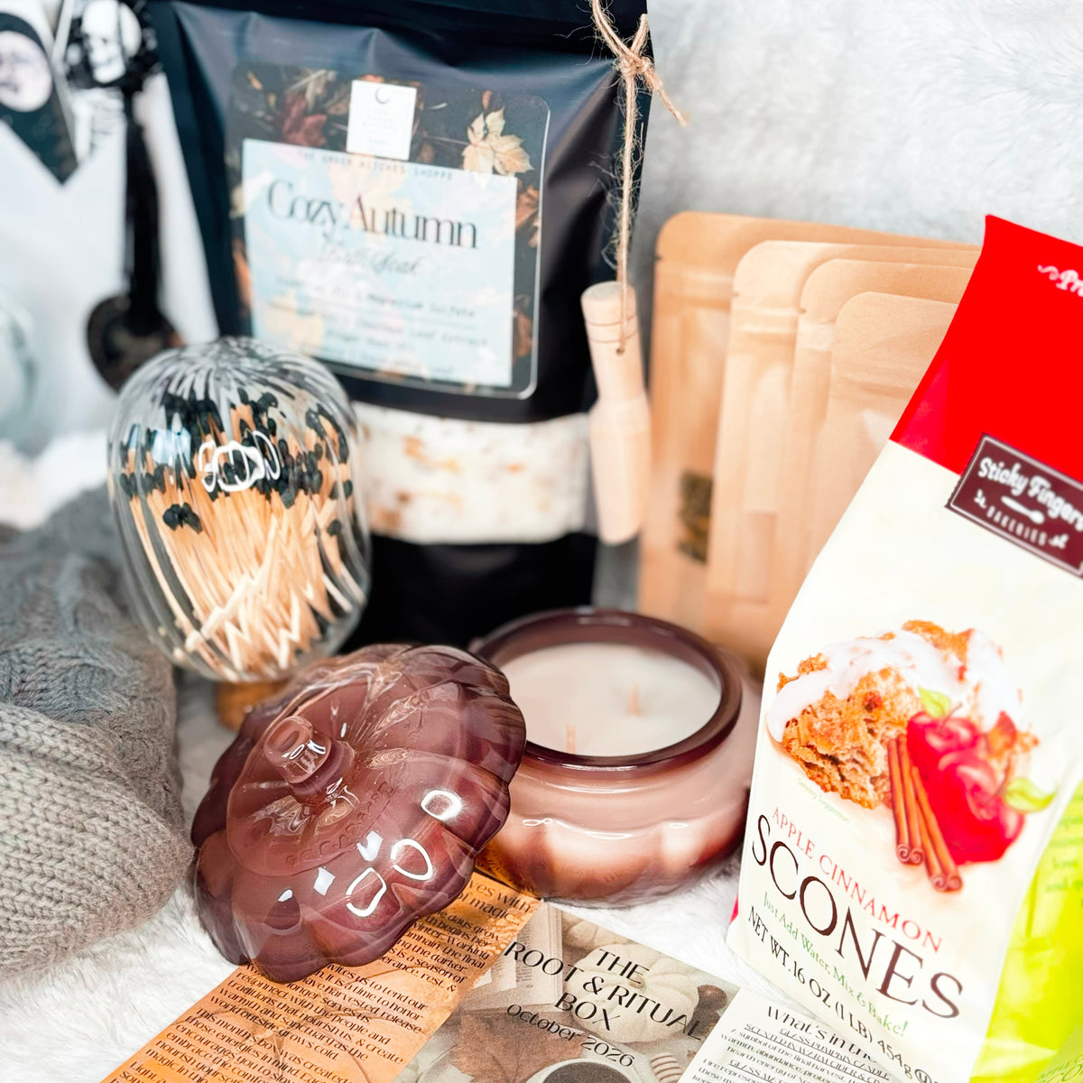 A cozy holiday gift set arranged on a white faux fur background, featuring gray cable-knit slipper socks, a glass mug and teapot with metal infuser, a bag of Cozy Autumn Bath Soak, a box of gourmet scone mix, a candle, and other warm seasonal accessories.