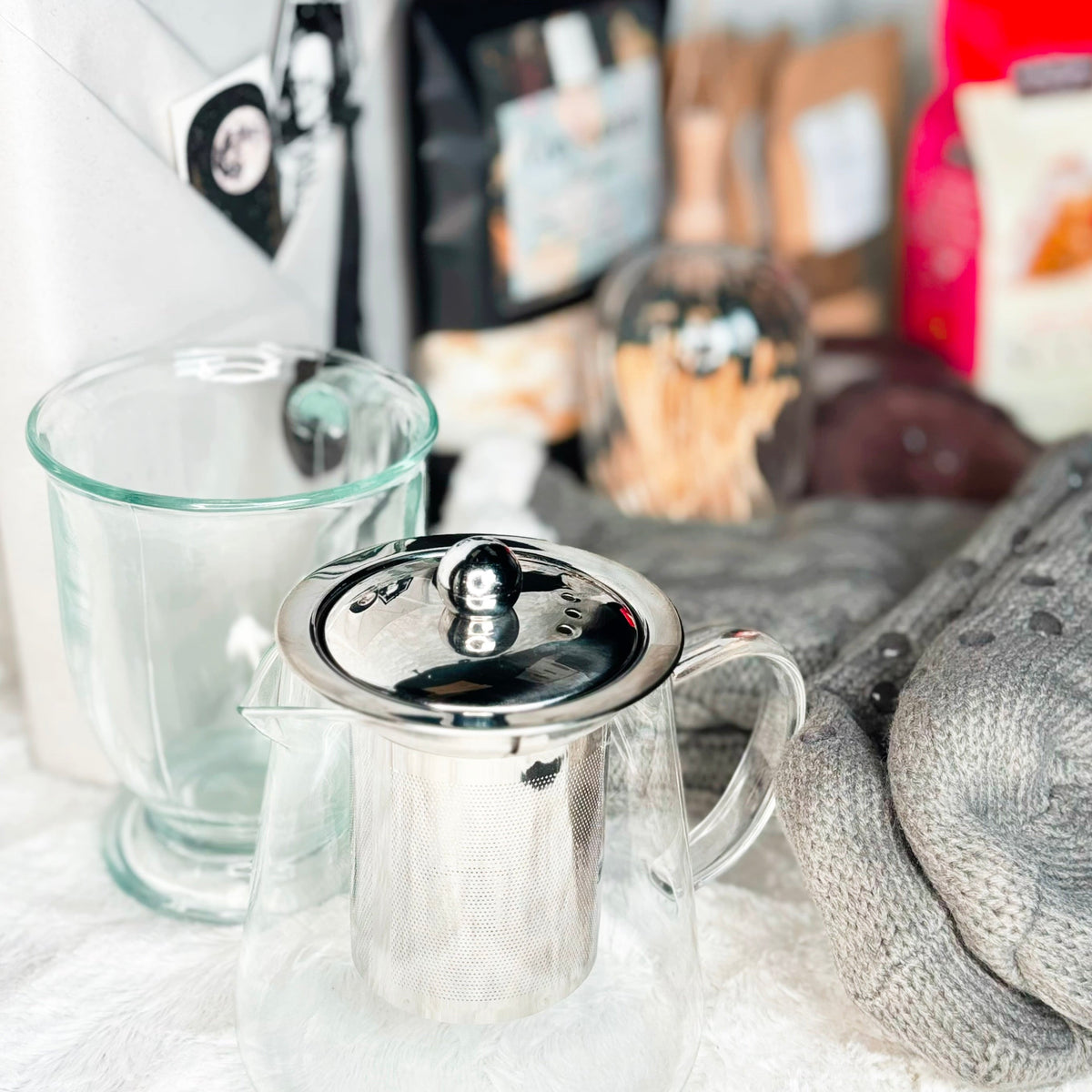A cozy holiday gift set arranged on a white faux fur background, featuring gray cable-knit slipper socks, a glass mug and teapot with metal infuser, a bag of Cozy Autumn Bath Soak, a box of gourmet scone mix, a candle, and other warm seasonal accessories.
