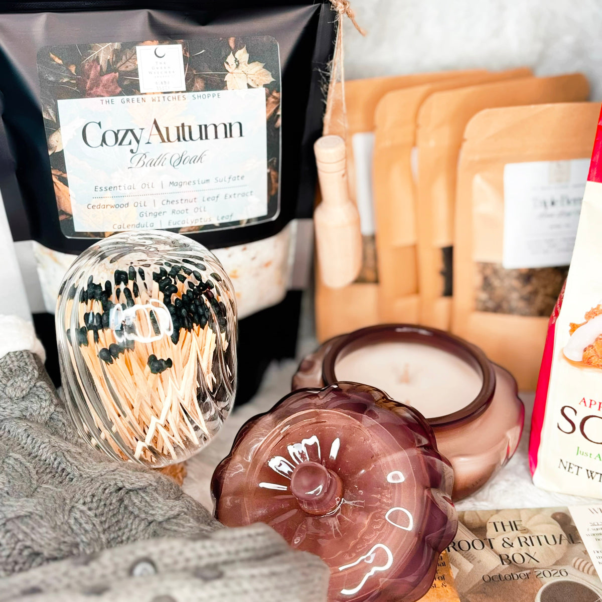 A cozy holiday gift set arranged on a white faux fur background, featuring gray cable-knit slipper socks, a glass mug and teapot with metal infuser, a bag of Cozy Autumn Bath Soak, a box of gourmet scone mix, a candle, and other warm seasonal accessories.